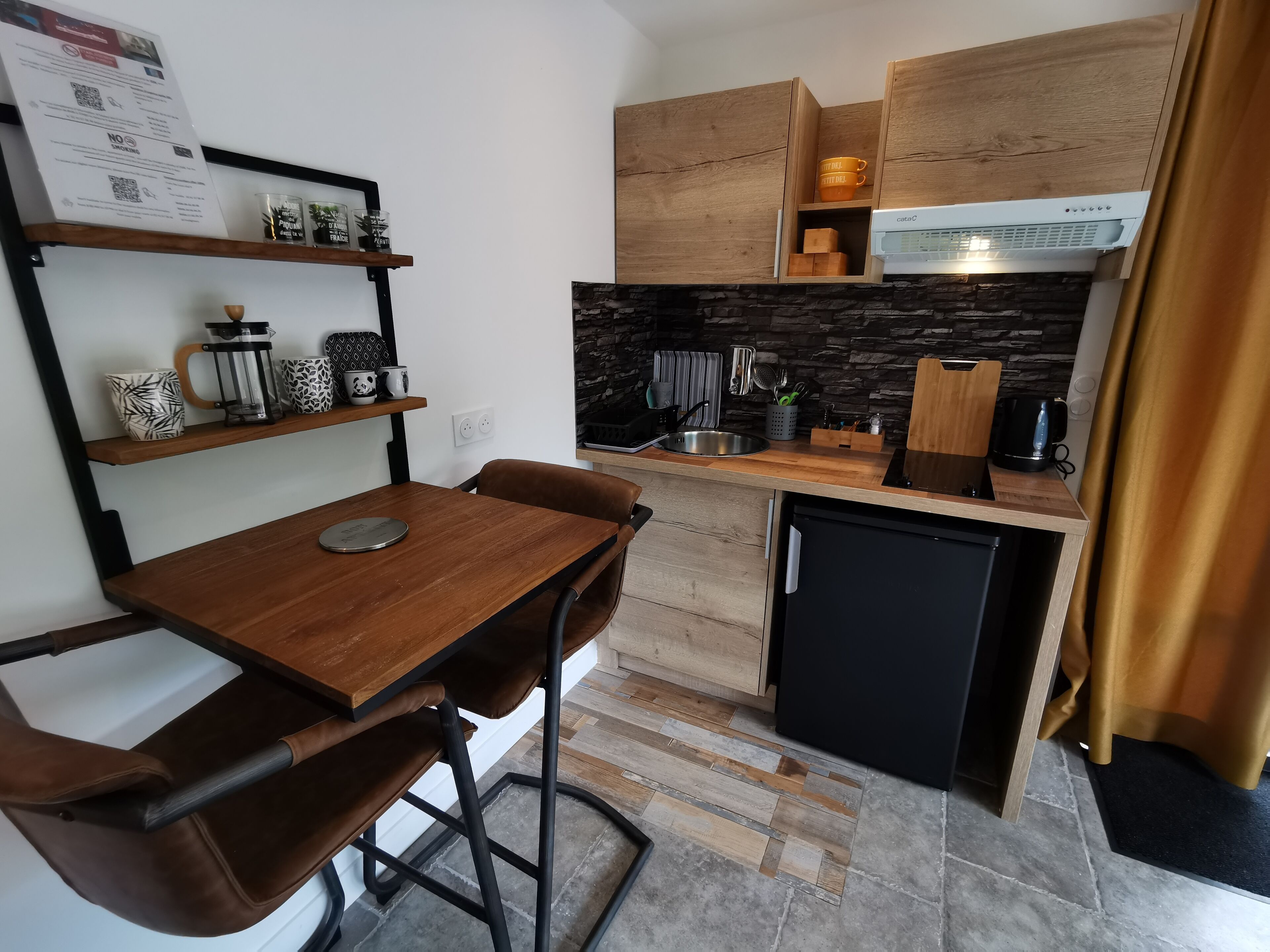 Une cuisine de taille réduite, avec des meubles de rangement en bois, un réfrigérateur noir et une machine à café posée sur le plan de travail.