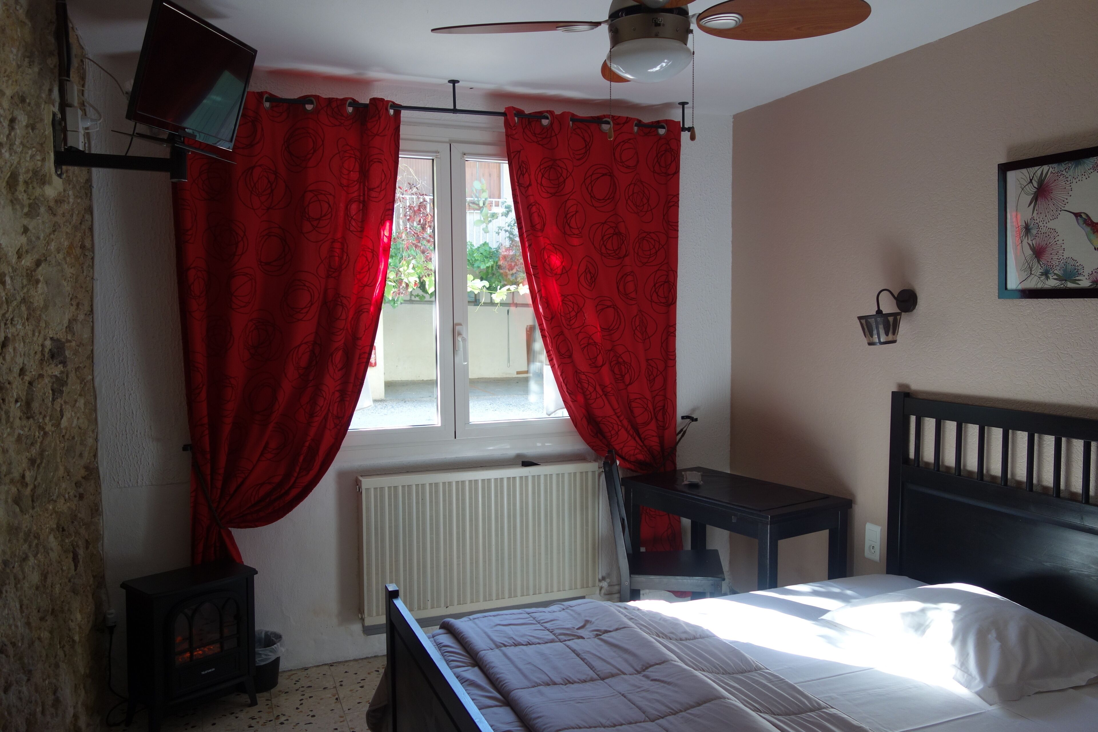 A bedroom with a bed, a heater, a TV mounted on the wall, and a window with red curtains.