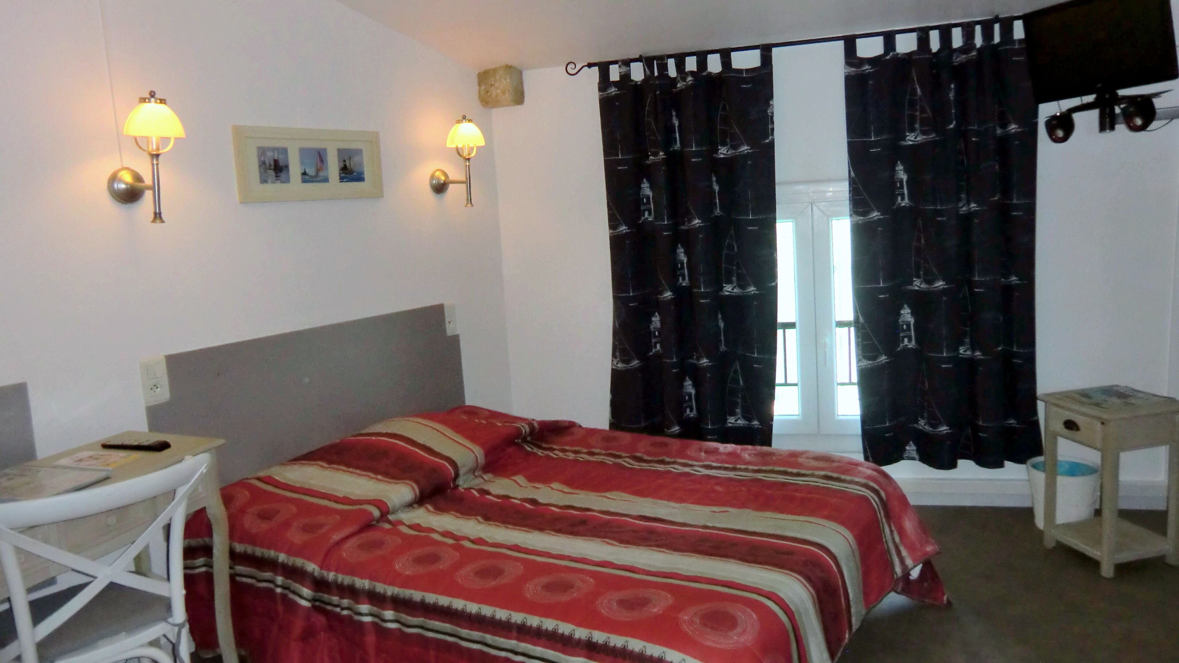 A room with a bed, two wall-mounted lamps, a desk with a chair, a small table, and a window with curtains.
