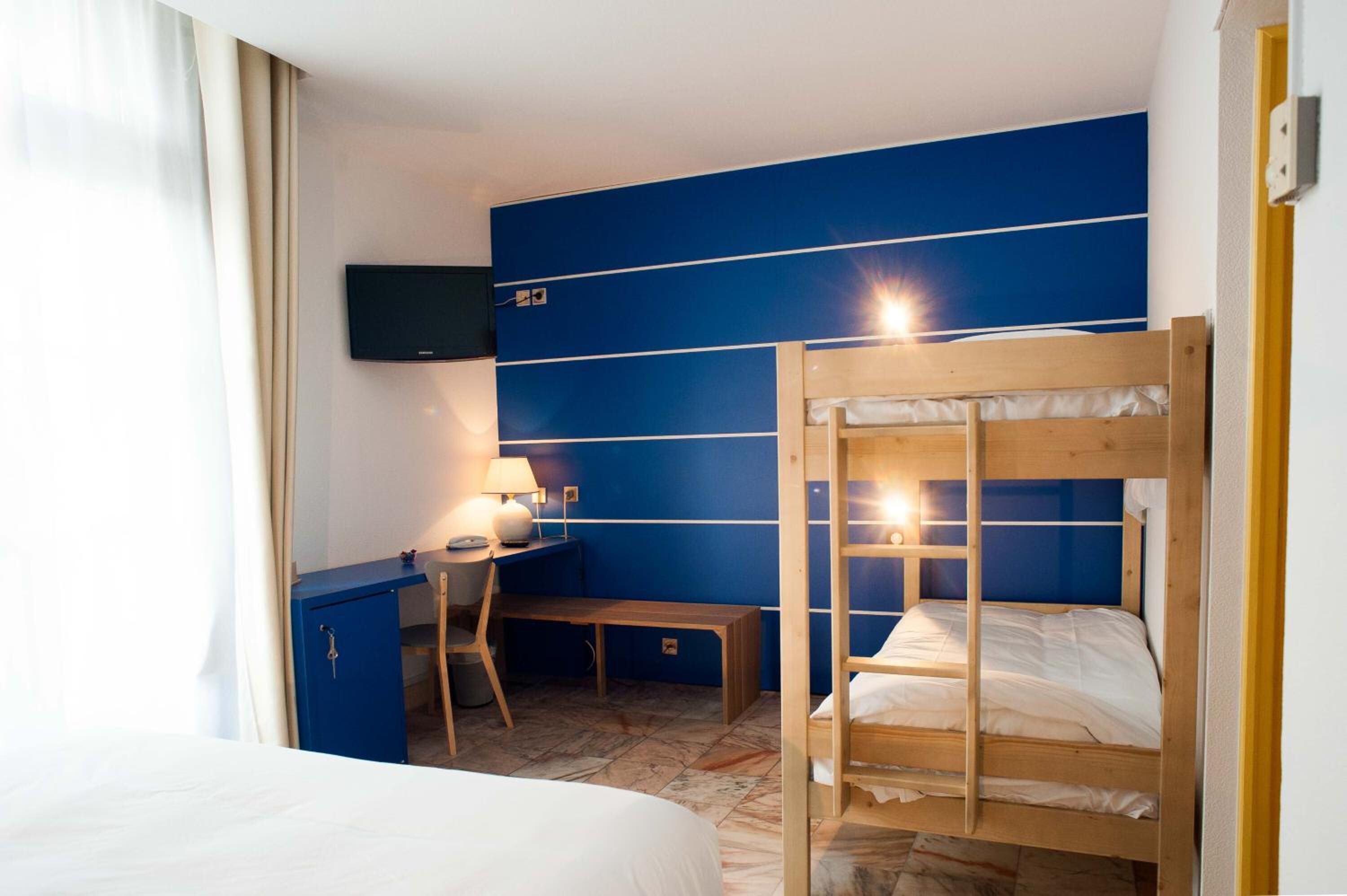 A room with a bunk bed, a desk with a chair, a television mounted on the wall, and a window with curtains.