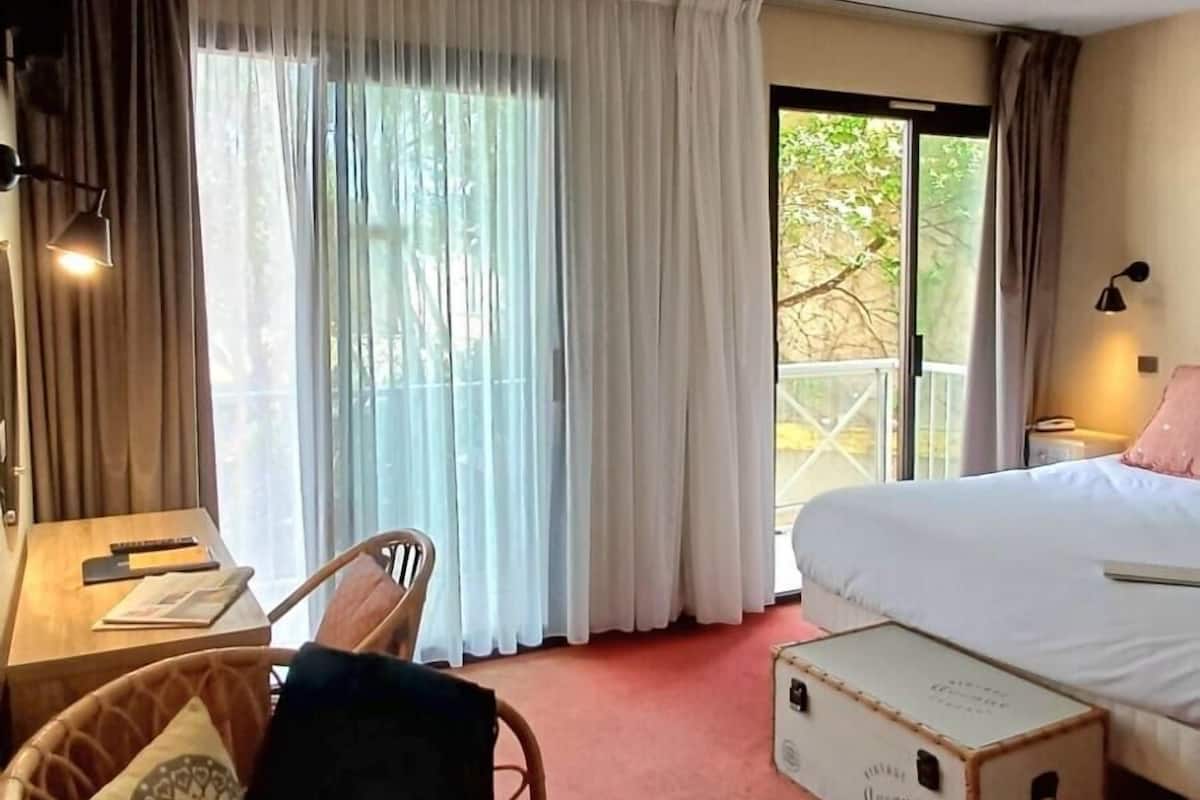 Een hotelkamer met een bed, een bureau met stoel, een raam met gordijnen en uitzicht op groen buiten.
