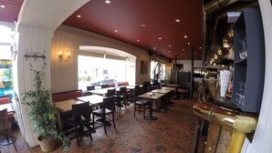 Un restaurante interior con mesas y sillas, una planta en maceta y una zona de bar con vasos y botellas.