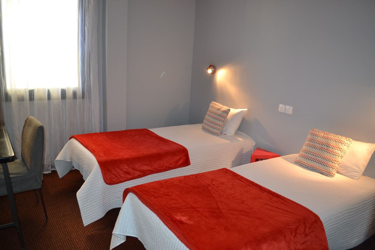 Een hotelkamer met twee bedden, elk met een rode deken en gestreepte kussens, een stoel, een bureau en een raam met gordijnen.