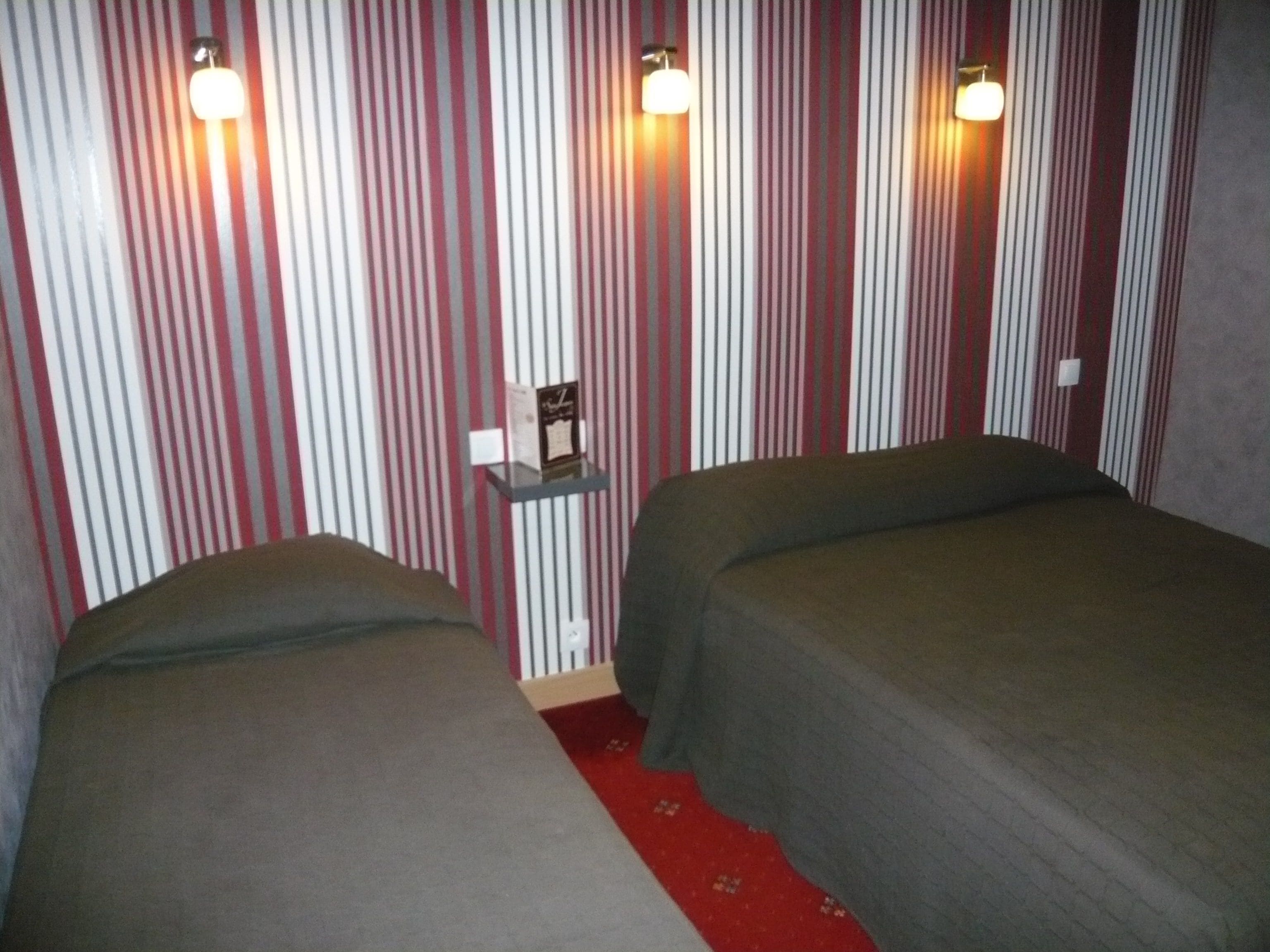 Een kamer met gestreept behang, twee bedden met grijze beddengoed, een klein plankje met een boek en twee wandlampen.