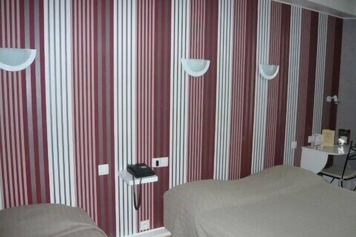 A hotel room with striped wallpaper, two beds, bedside tables, and a small desk with a lamp.