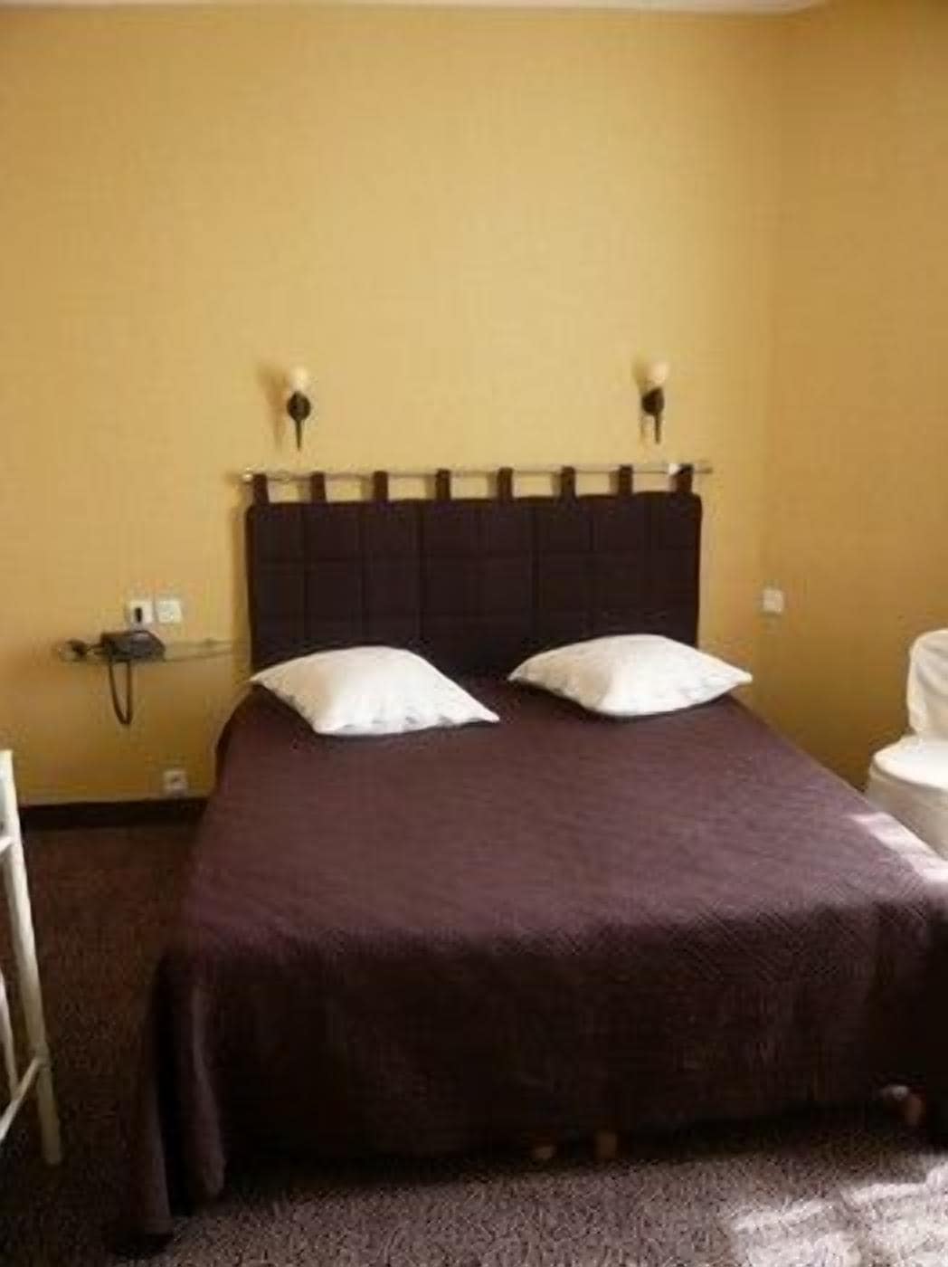 Uma cama de solteiro com cabeceira escura, duas almofadas e um cobre-leito marrom em um quarto com paredes amarelas.