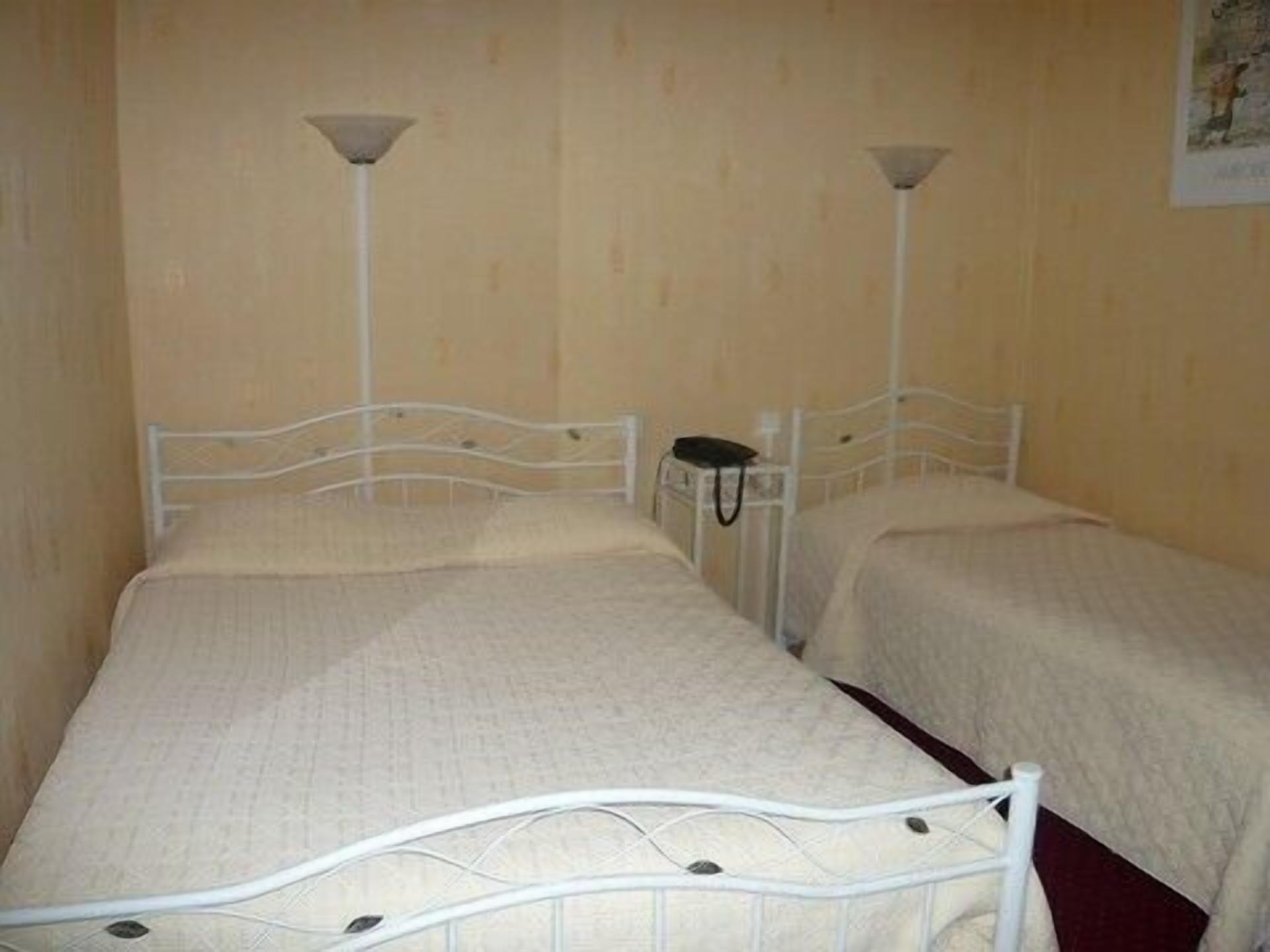 A room with two single beds, each with a white metal frame and a headboard. There is a small bedside table with a lamp on each bed. The walls are beige, and there is a framed picture on the wall.