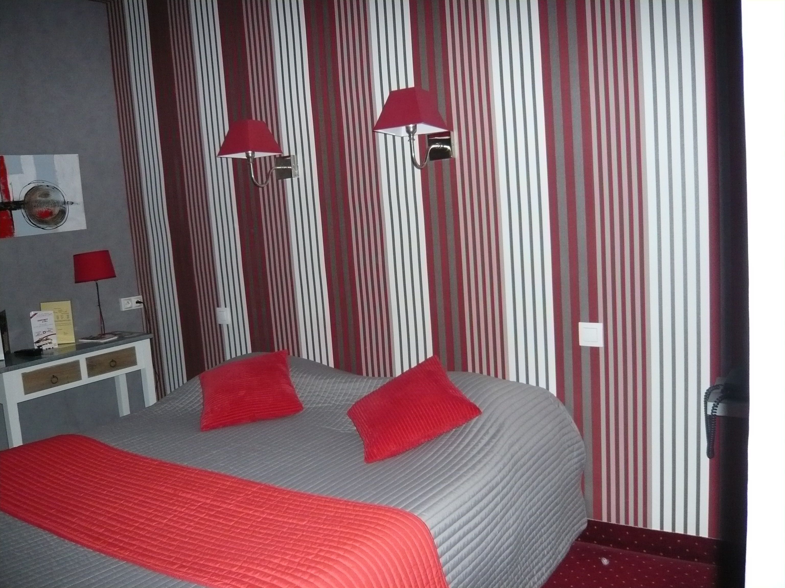 A hotel room with a bed, two red lamps, a white nightstand, and a striped wall.