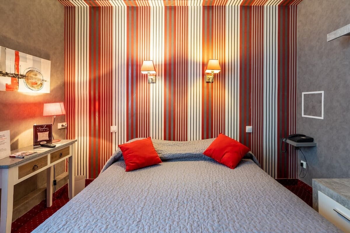 A hotel room with a large bed, two red pillows, a desk with a lamp, and a telephone.