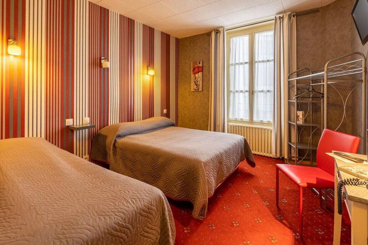 A hotel room with two beds, a red chair, a desk, a radiator, and a window with curtains.
