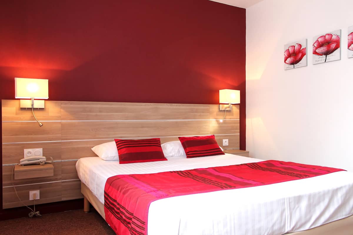 A hotel room with a wooden headboard, a white bed with red and black pillows, two bedside lamps, a nightstand with a phone, and a wall with three red flower paintings.