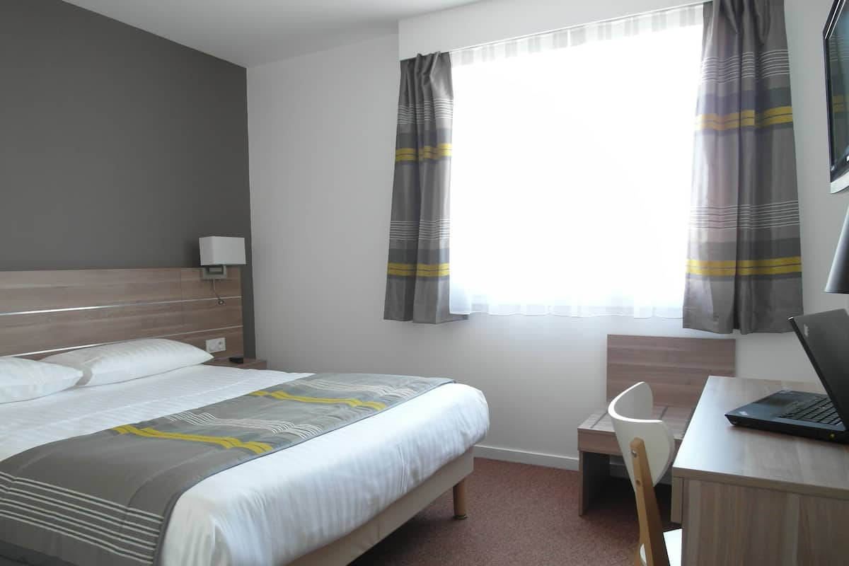 A hotel room with a bed, a desk with a laptop, a chair, a nightstand, and a window with curtains.