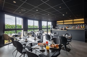 Uma área de jantar moderna com mesas preparadas para o café da manhã, amplas janelas com vista para áreas verdes e um horizonte urbano, e um bar com diversas bebidas.