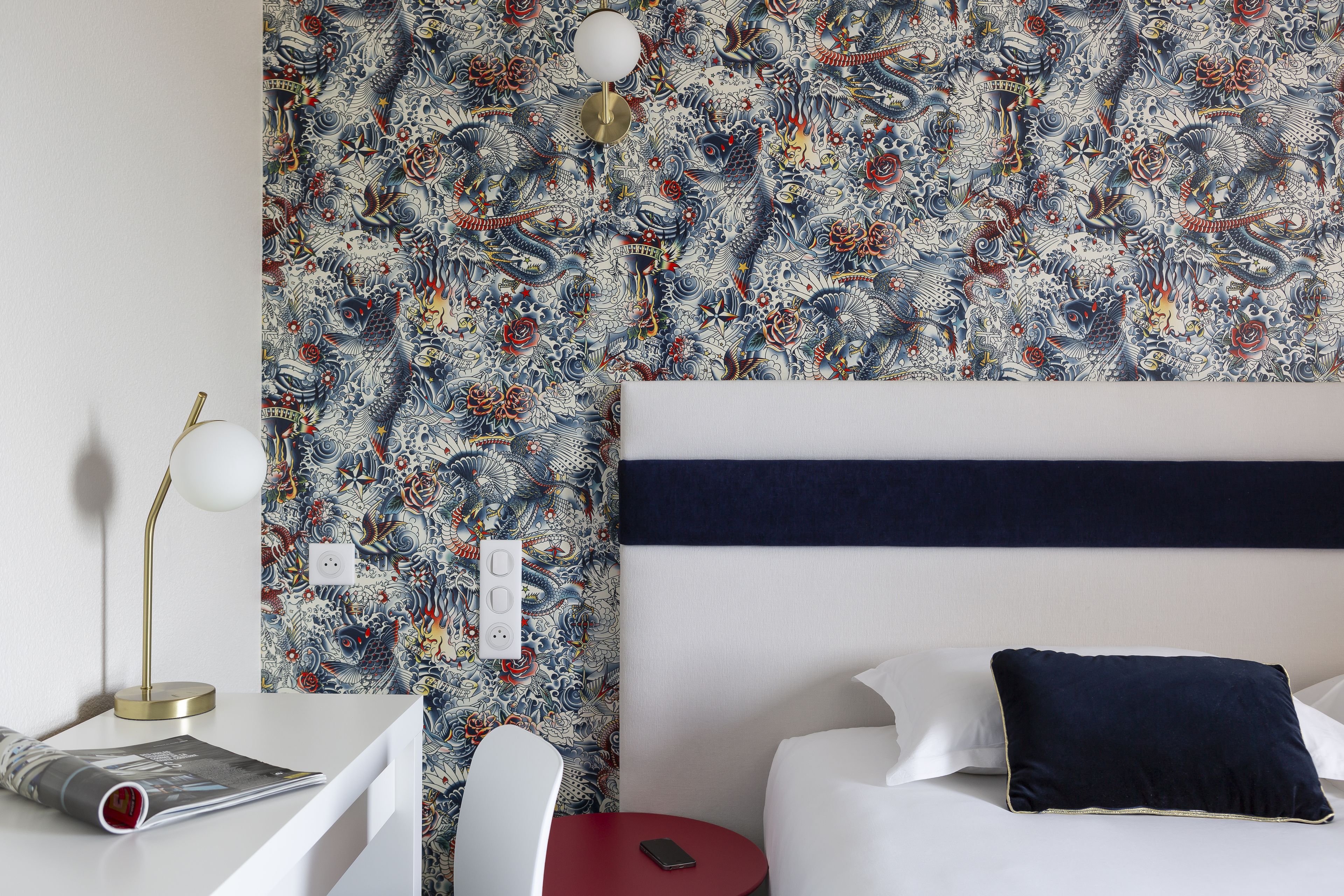 A hotel room with a patterned wallpaper, a white bed with a navy pillow, a red side table, a white chair, and a desk with a lamp and a magazine.
