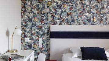 A hotel room with a patterned wallpaper, a white bed with a navy pillow, a red side table, a white chair, and a desk with a lamp and a magazine.