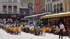 Un café al aire libre concurrido, con sillas y mesas amarillas, rodeado de edificios coloridos con varios letreros y una gran escultura azul.