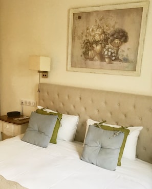 Habitación de hotel con una cama grande, dos almohadas, una mesita de noche, una lámpara y un cuadro con flores en la pared.