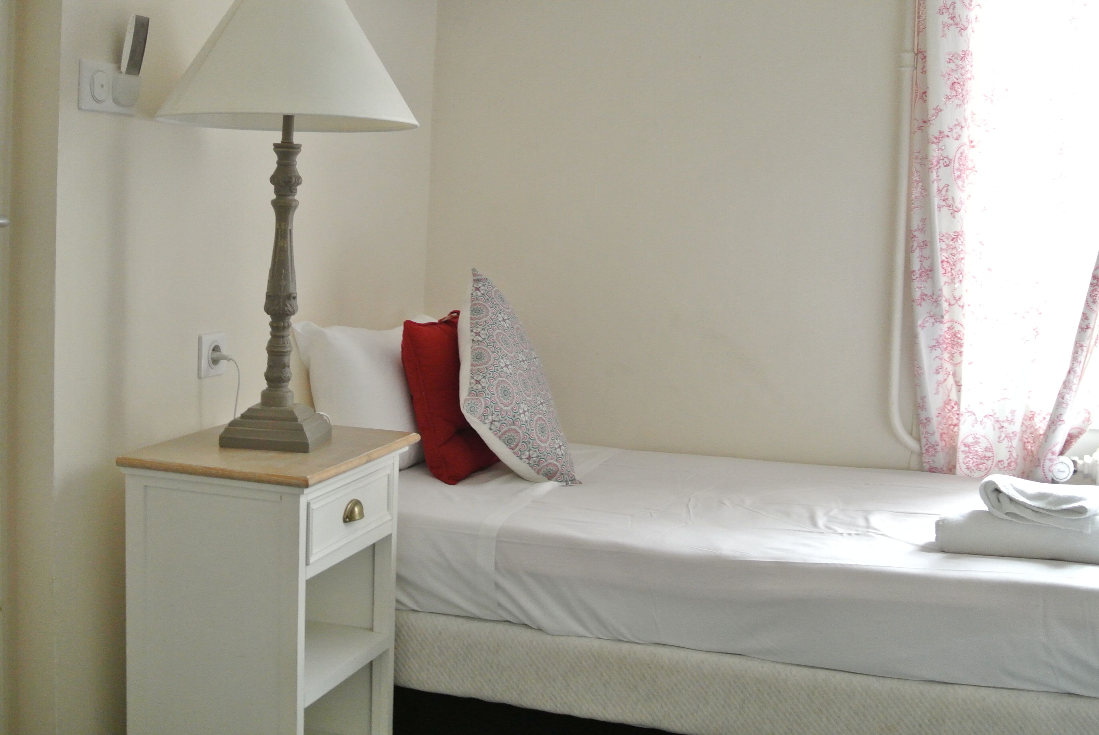 A neatly made bed with white bedding, a bedside lamp, a red pillow, and a patterned pillow on a white nightstand.