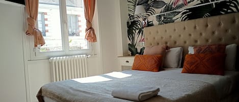 A bedroom with a bed, a window with curtains, a radiator, and a wall with a tropical wallpaper.