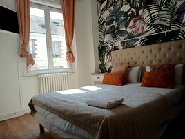 Une chambre à coucher avec un lit, une fenêtre avec des rideaux, un radiateur et un mur recouvert d’un papier peint à motifs tropicaux.