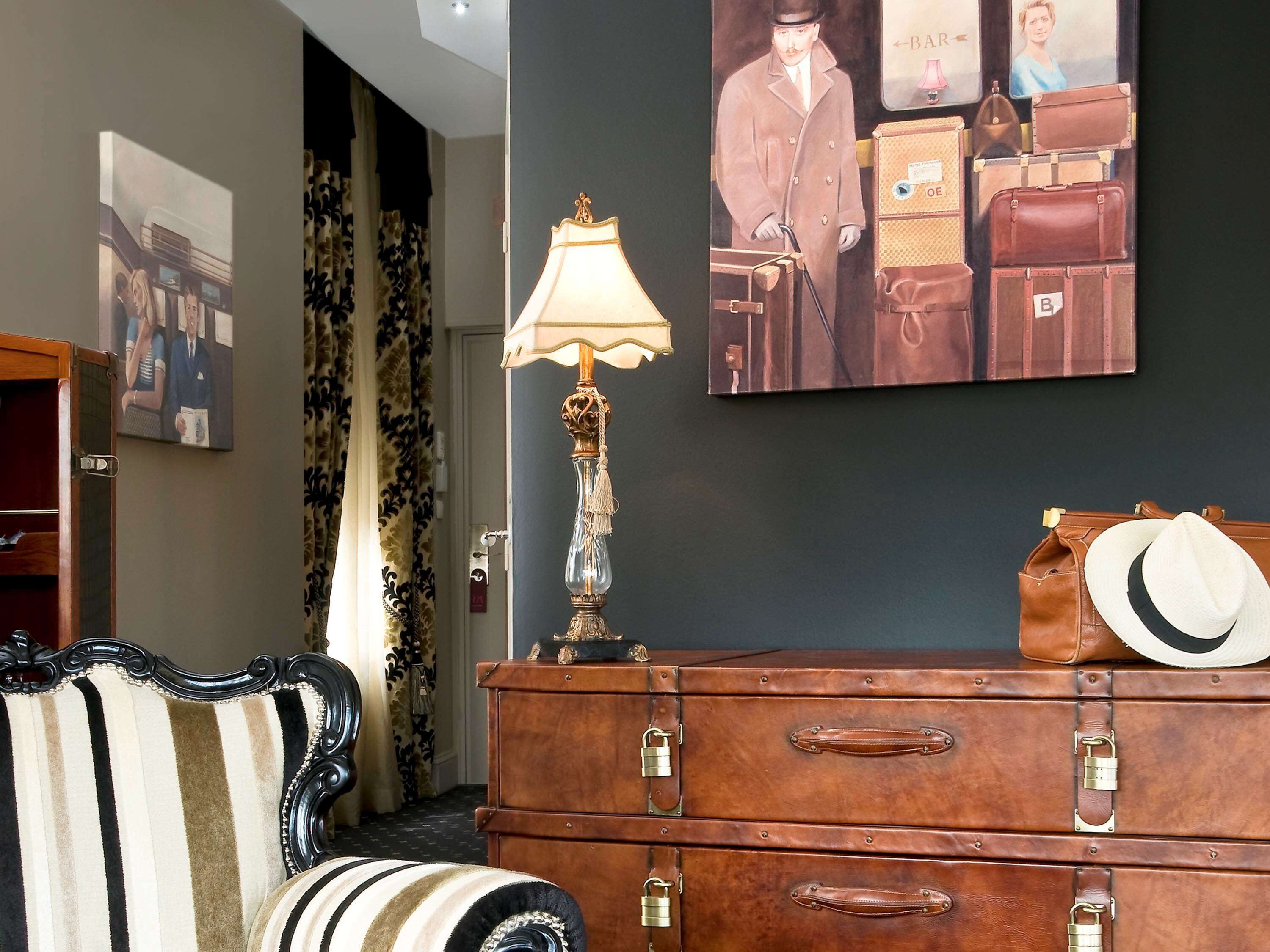 Quarto com poltrona listrada, luminária, cômoda com malas e pintura de um homem carregando bagagem.