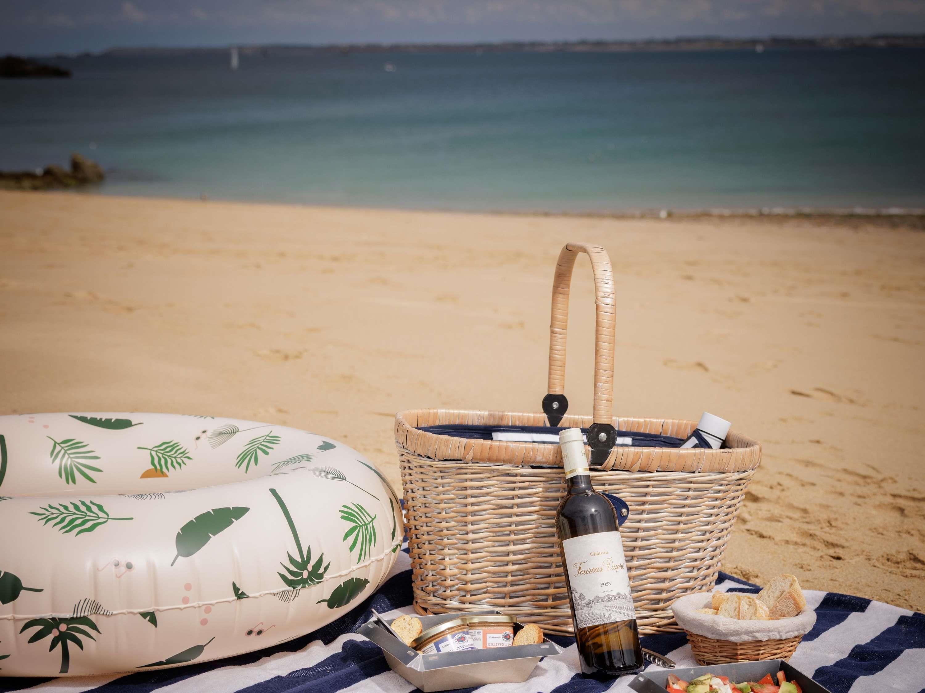 A beach scene with a wicker basket, a bottle, and food items on a blanket.