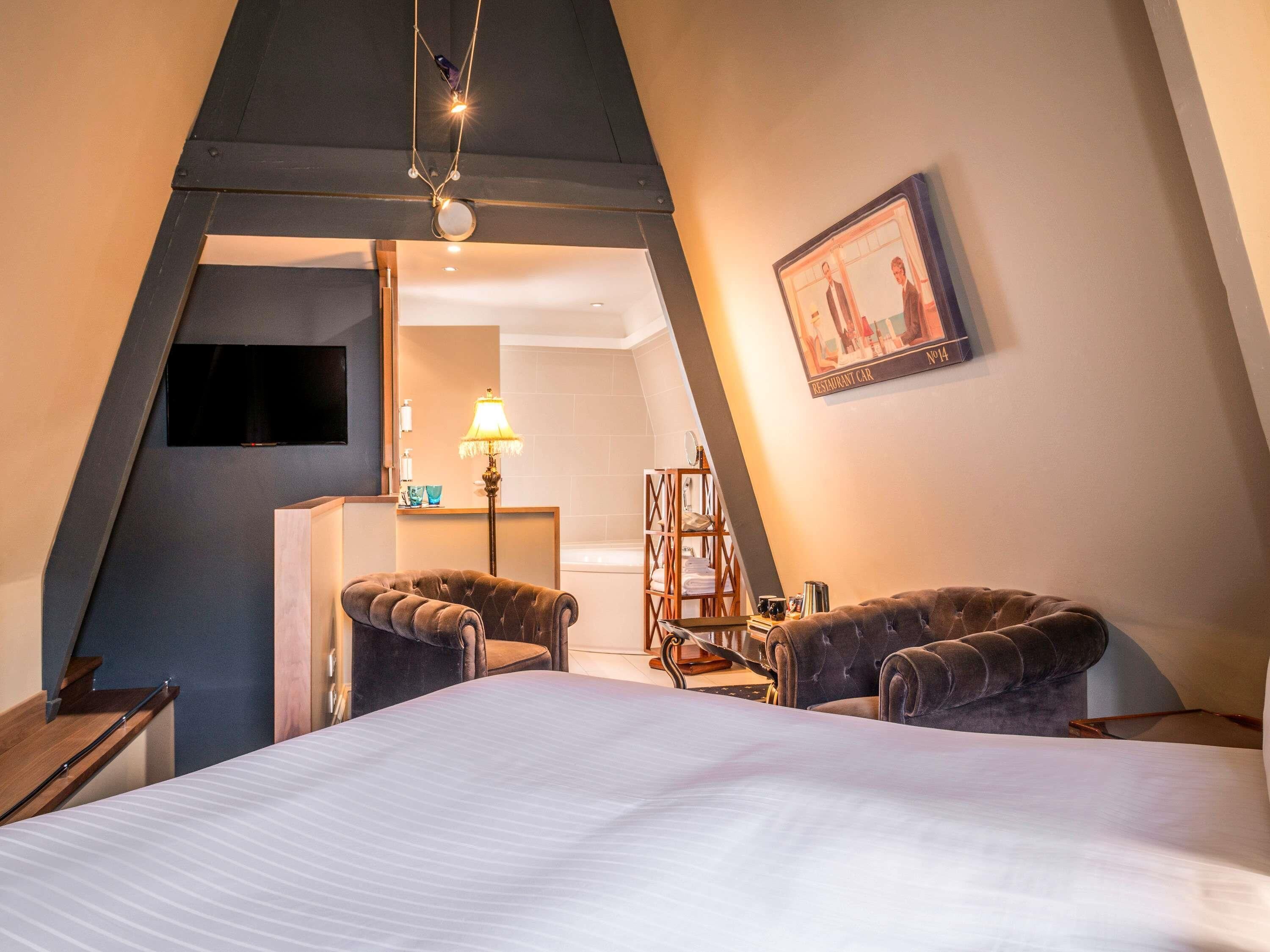 Une pièce triangulaire comprenant un lit, deux fauteuils, un bureau, une lampe et un tableau accroché au mur.