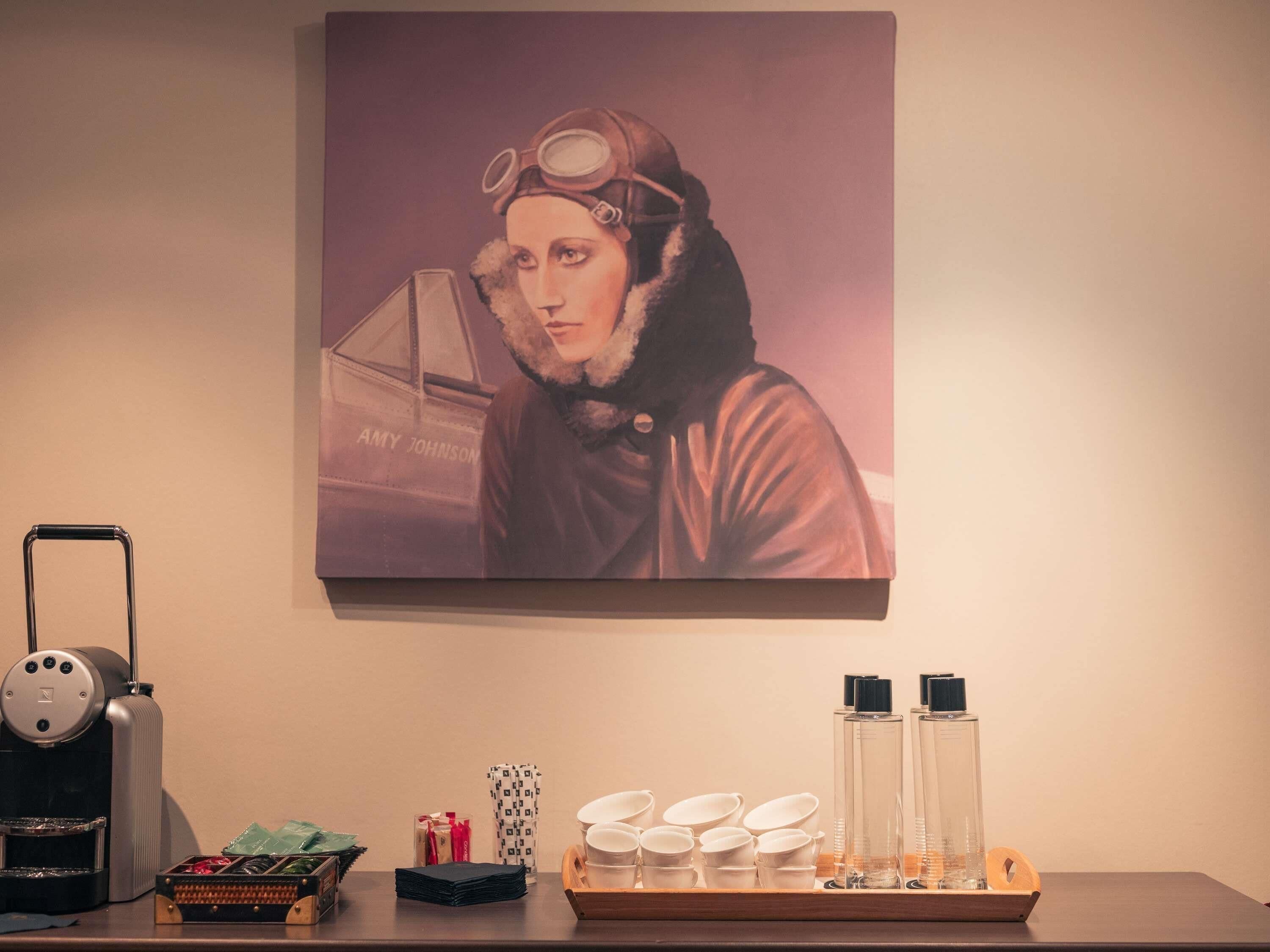 Un quadro appeso alla parete raffigura una persona con occhiali protettivi, una macchina del caffè e un vassoio con tazze e bottiglie.