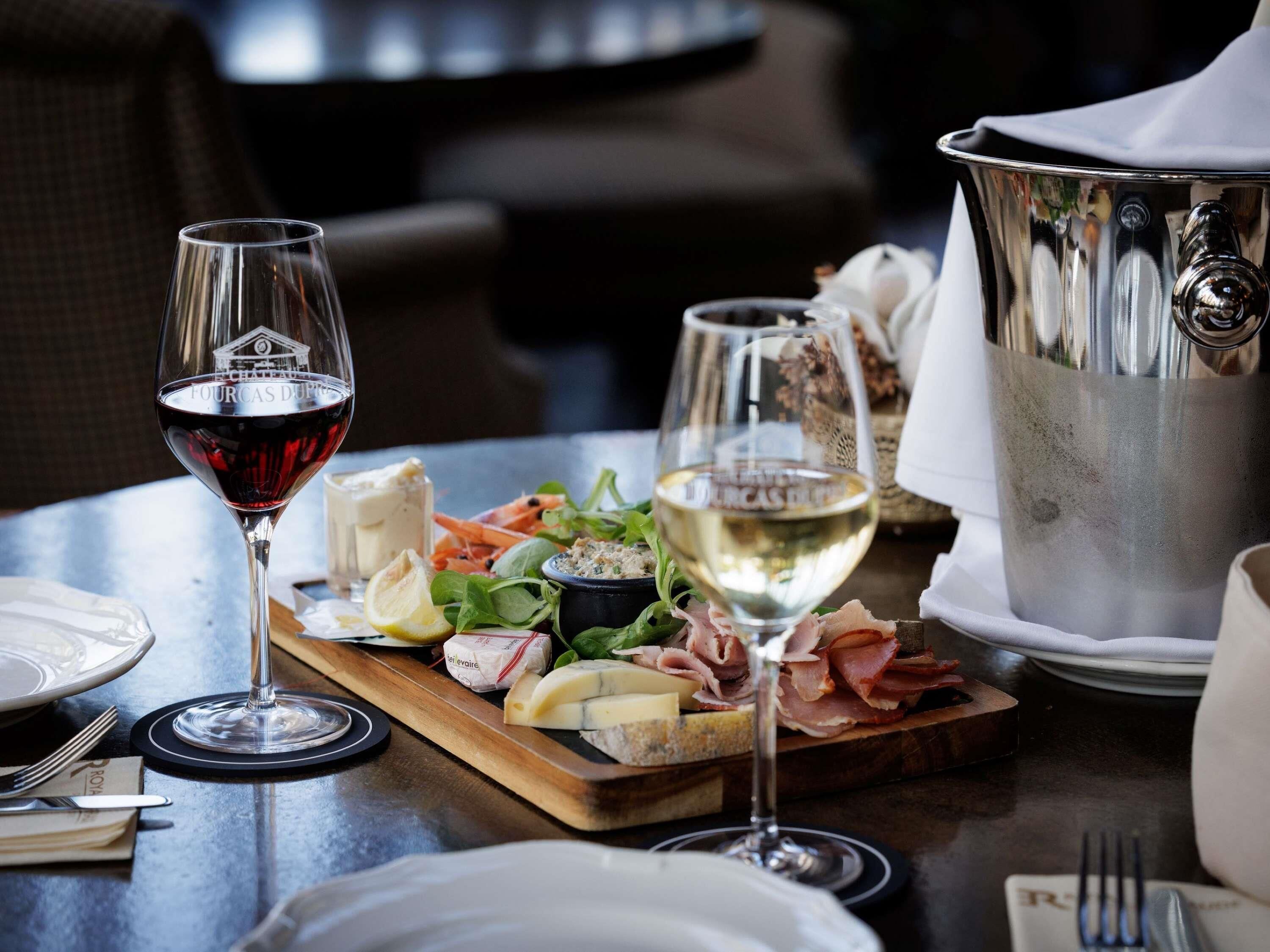 A dining table with a wine glass, a cheese board with various cheeses and meats, a pitcher, and a bottle of white wine.