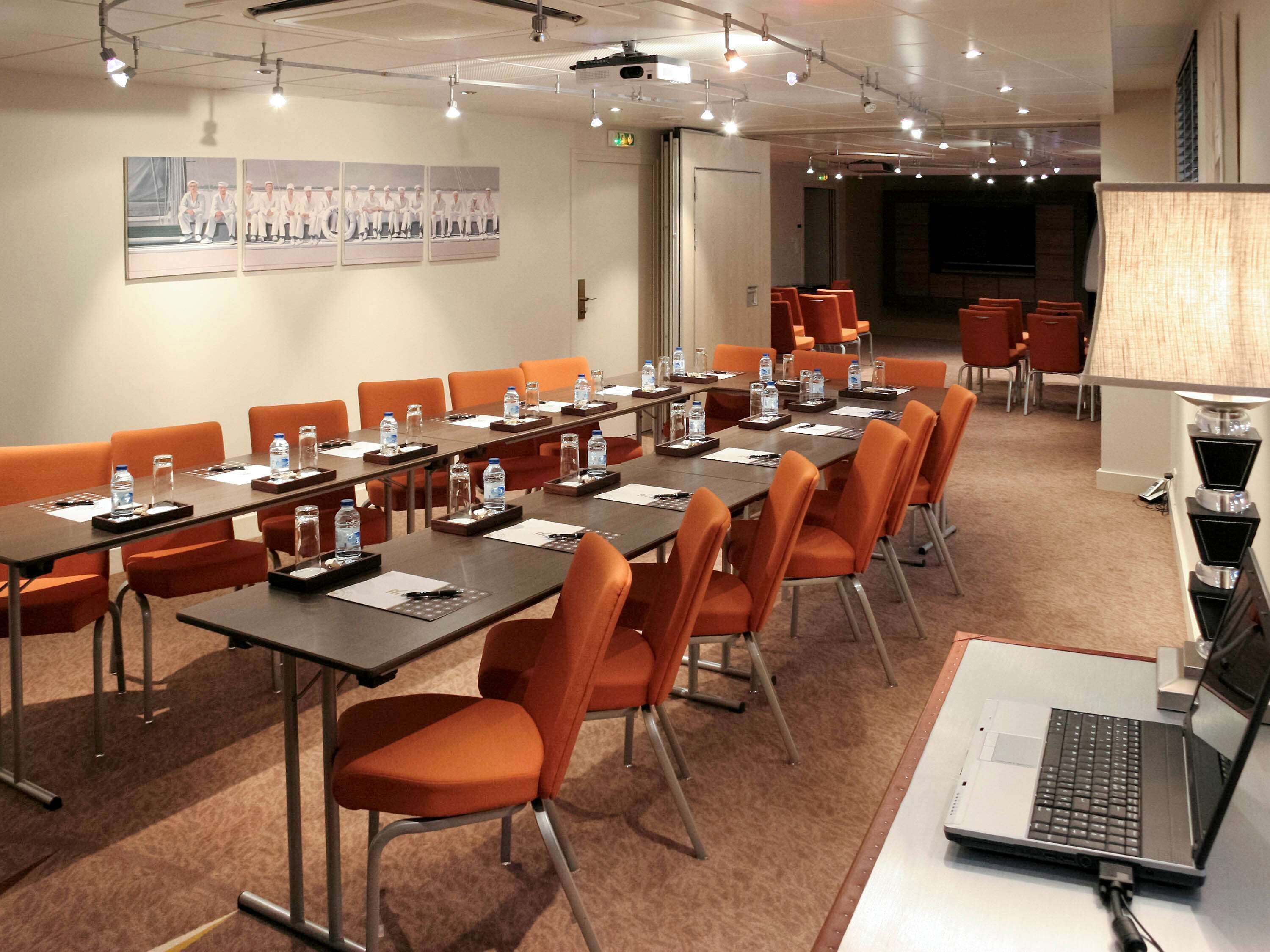 Un salón de conferencias con largas mesas, sillas anaranjadas, botellas de agua y un ordenador portátil sobre un escritorio.