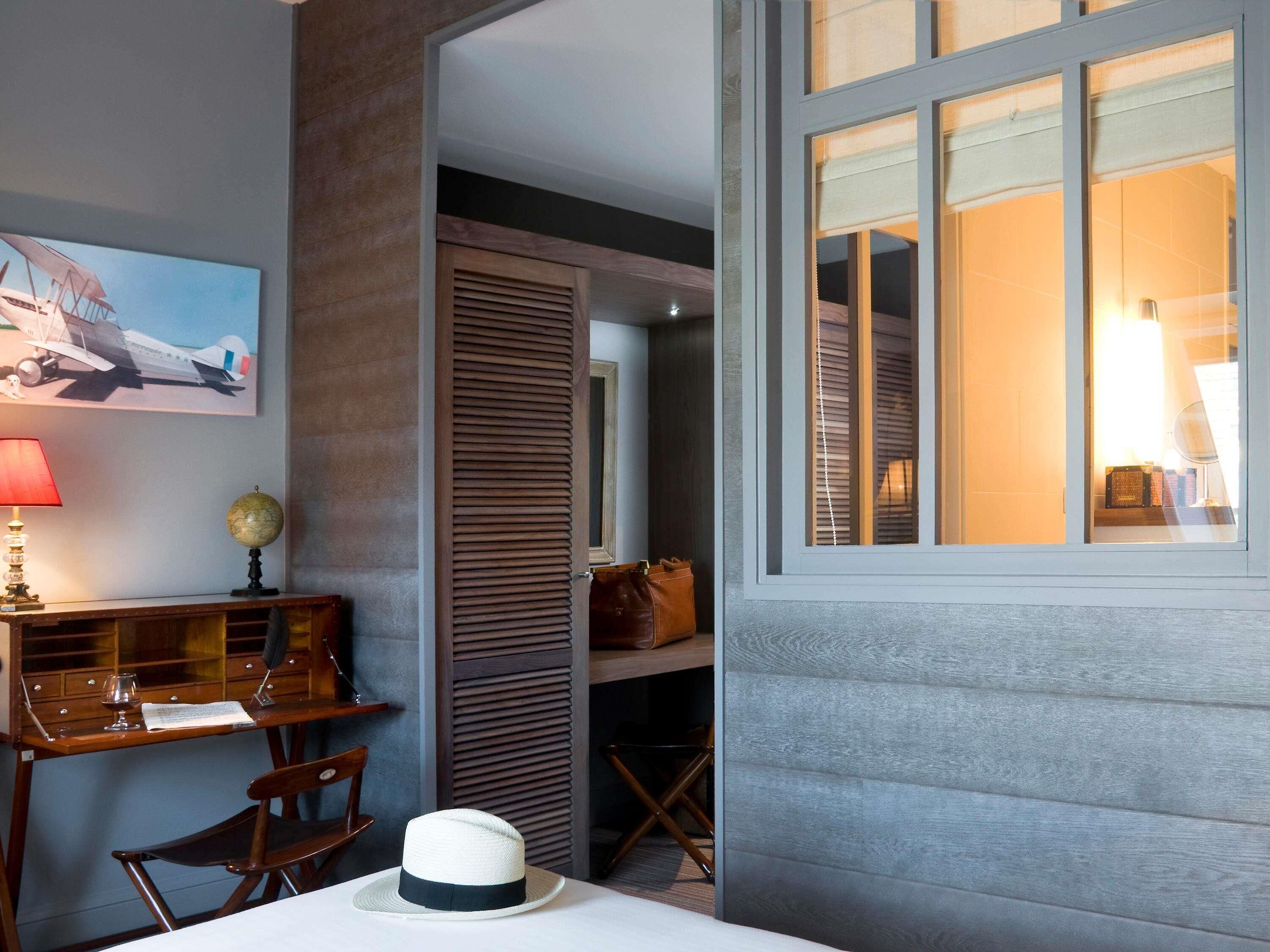 A room with a desk, chair, and a hat on the table. There is a window with blinds, a lamp, and a globe on the desk.