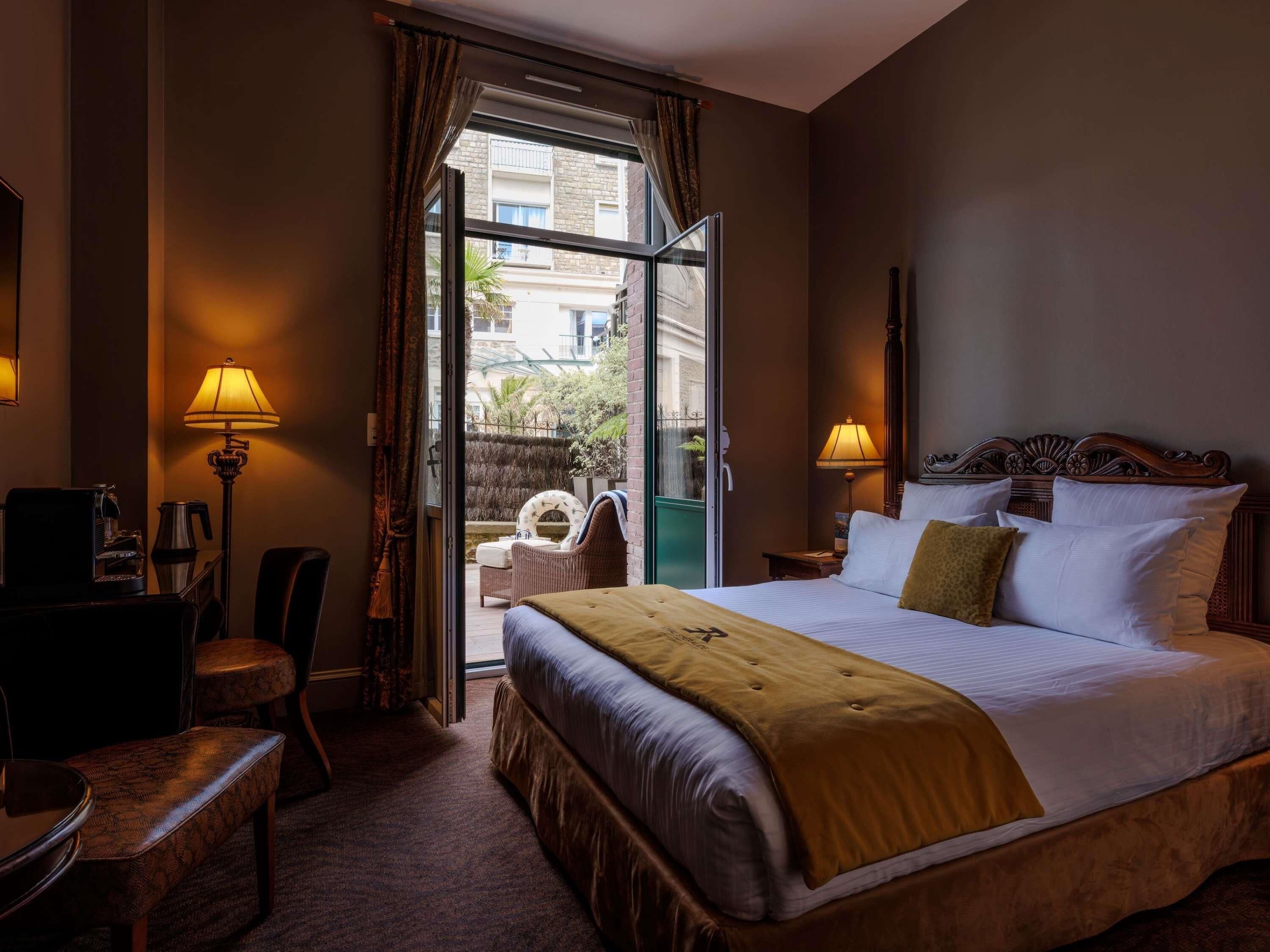 Een hotelkamer met een groot bed, een bureau met een lamp, een stoel en uitzicht door een glazen deur.