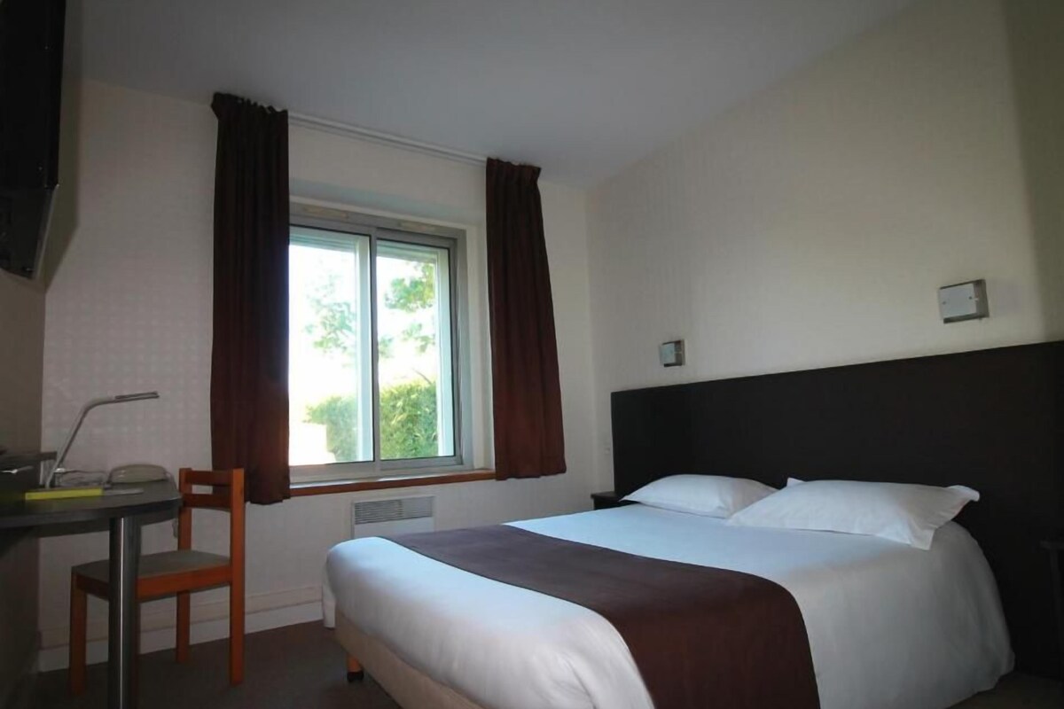 A hotel room with a bed, a desk, a chair, a window with curtains, and a thermostat on the wall.