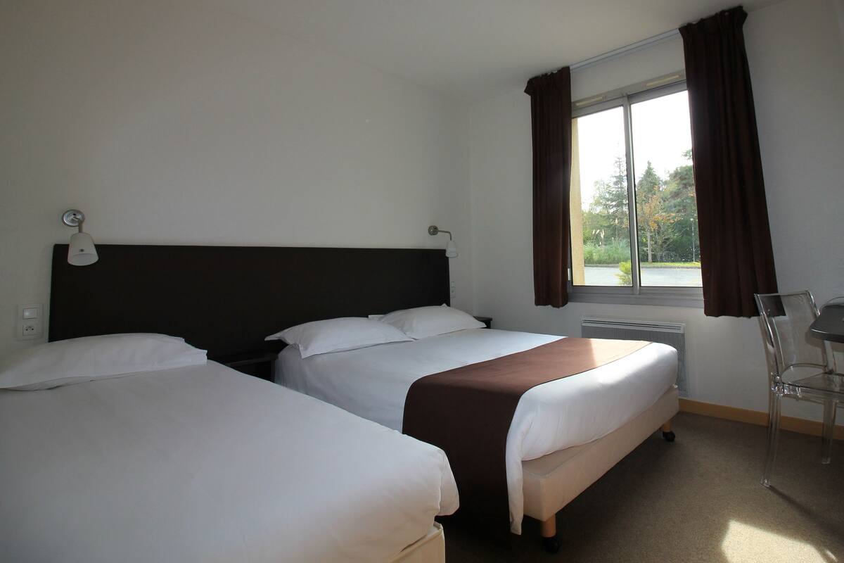 A hotel room with two beds, a window with curtains, a desk with a chair, and a lamp.