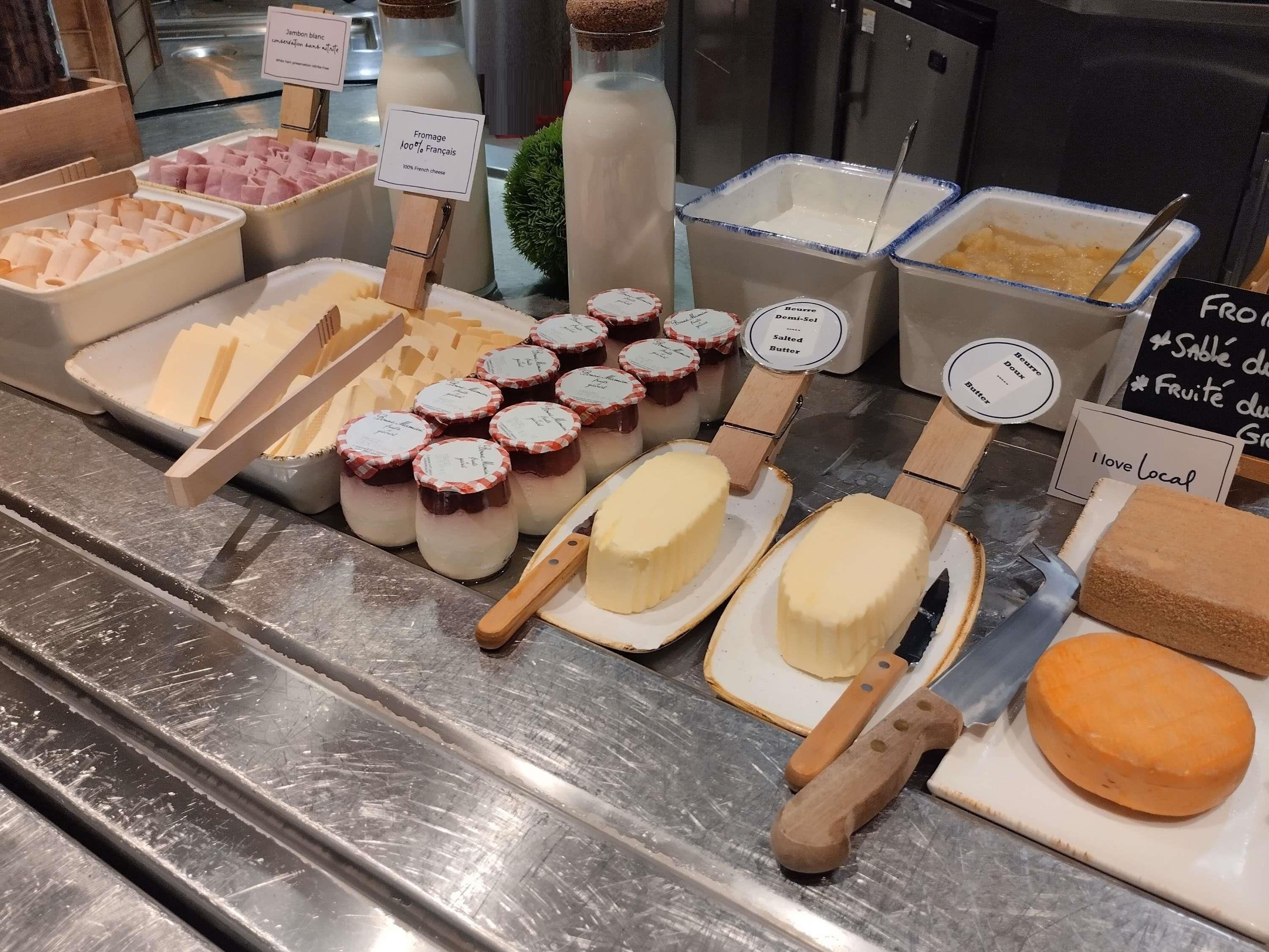 Diversos produtos lácteos, como queijo, manteiga e iogurte, expostos em um balcão de metal com etiquetas e placas de preço.