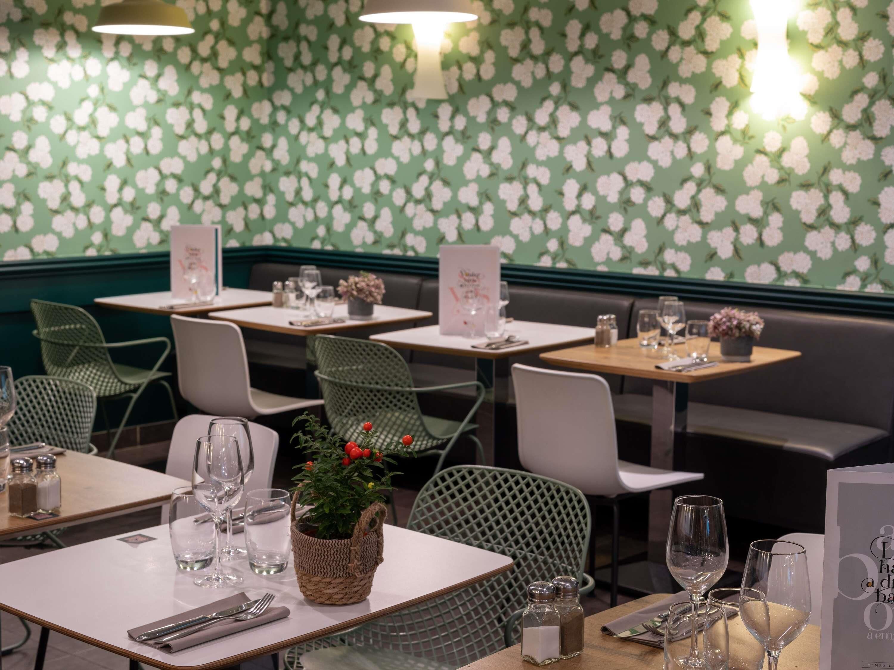 A restaurant interior with green floral wallpaper, green chairs, tables set with wine glasses, and a menu stand.