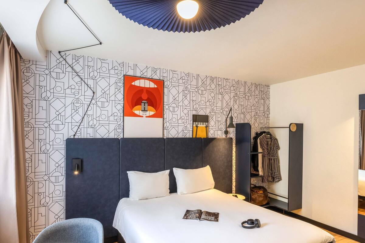A modern hotel room with a geometric patterned wallpaper, a large bed with white bedding, a bedside table with a magazine and headphones, a chair, and a wardrobe with hanging clothes.