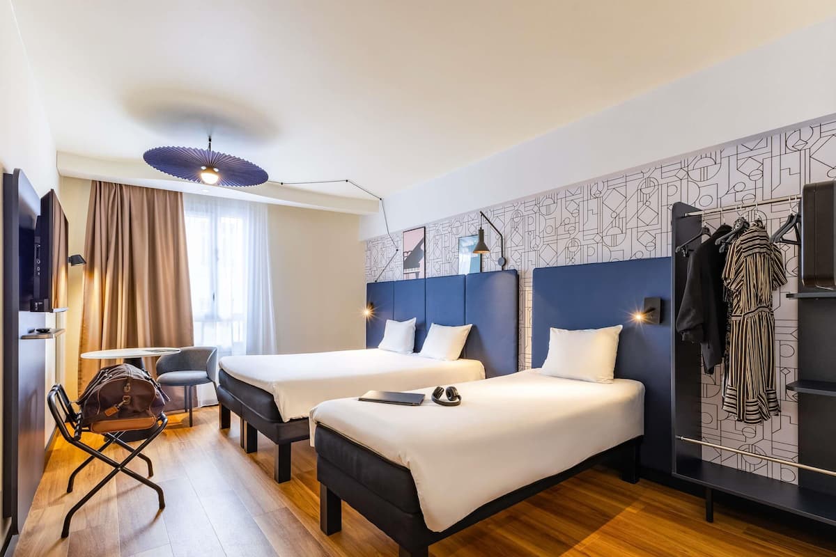 A hotel room with two beds, a desk, a chair, a wardrobe, and a ceiling fan.