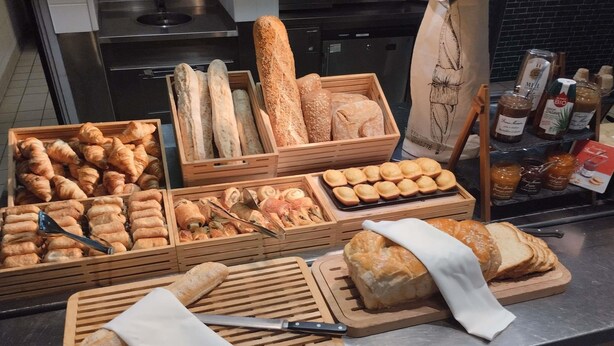 A variety of breads and pastries displayed in wooden crates and wooden boards, with a knife and a pair of tongs on the counter.