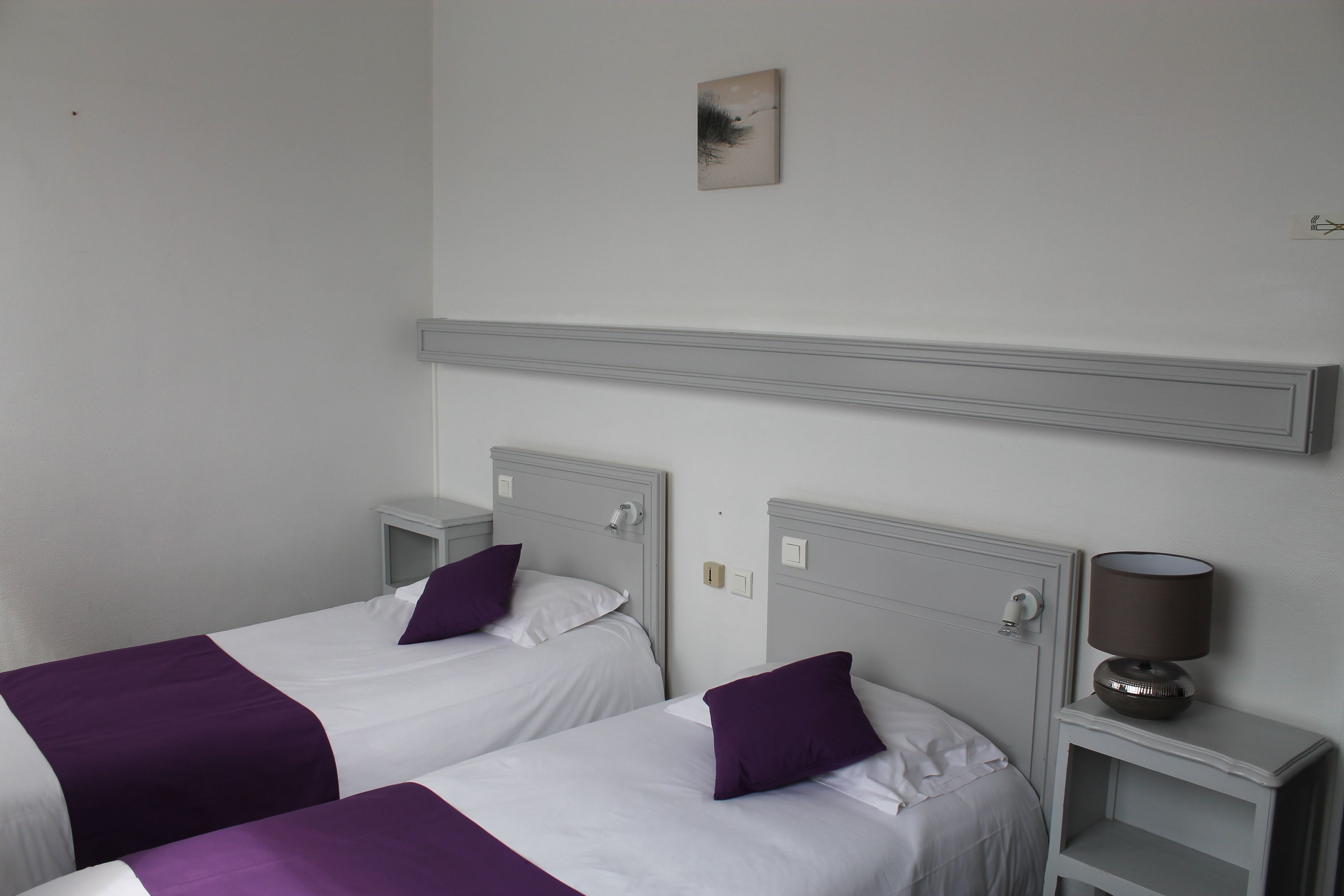 Une chambre d’hôtel avec deux lits, chacun doté d’une tête de lit blanche et d’une literie violette. Il y a une table de chevet avec une lampe d’un côté et une petite table avec une lampe de l’autre.