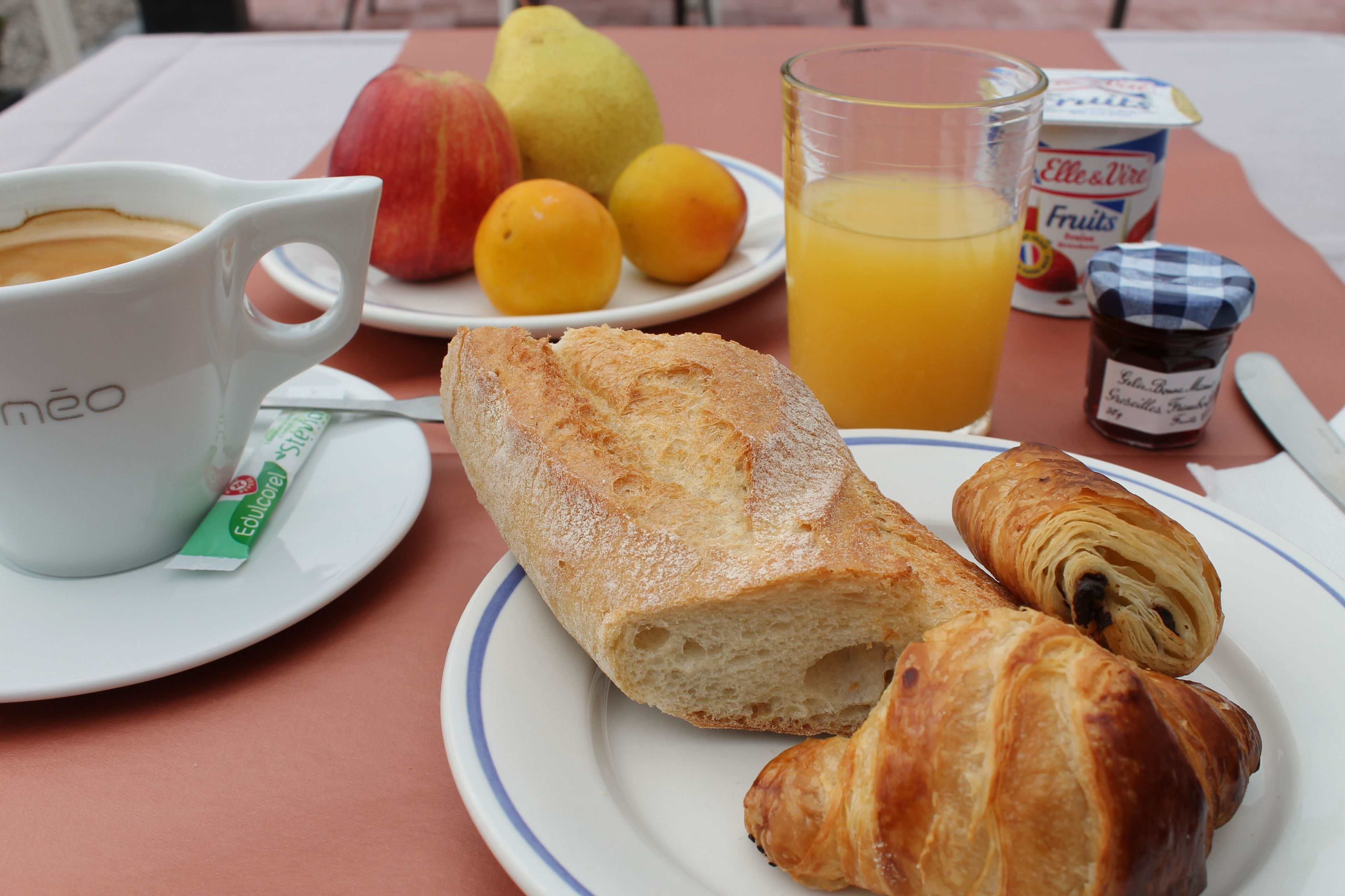 A breakfast setting with a croissant, a glass of orange juice, a plate of fruit, a coffee cup, and a jar of jam.