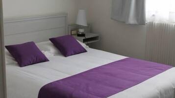 A bed with white and purple bedding, a nightstand with a lamp, and a window with curtains.