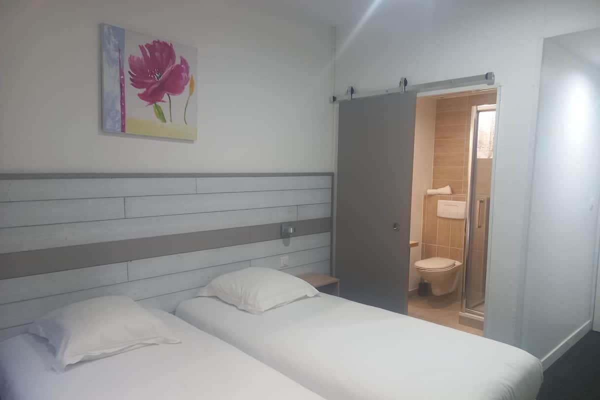 Une chambre d’hôtel avec deux lits simples, une tête de lit en bois, un tableau représentant des fleurs et une salle de bain visible à travers une porte ouverte.