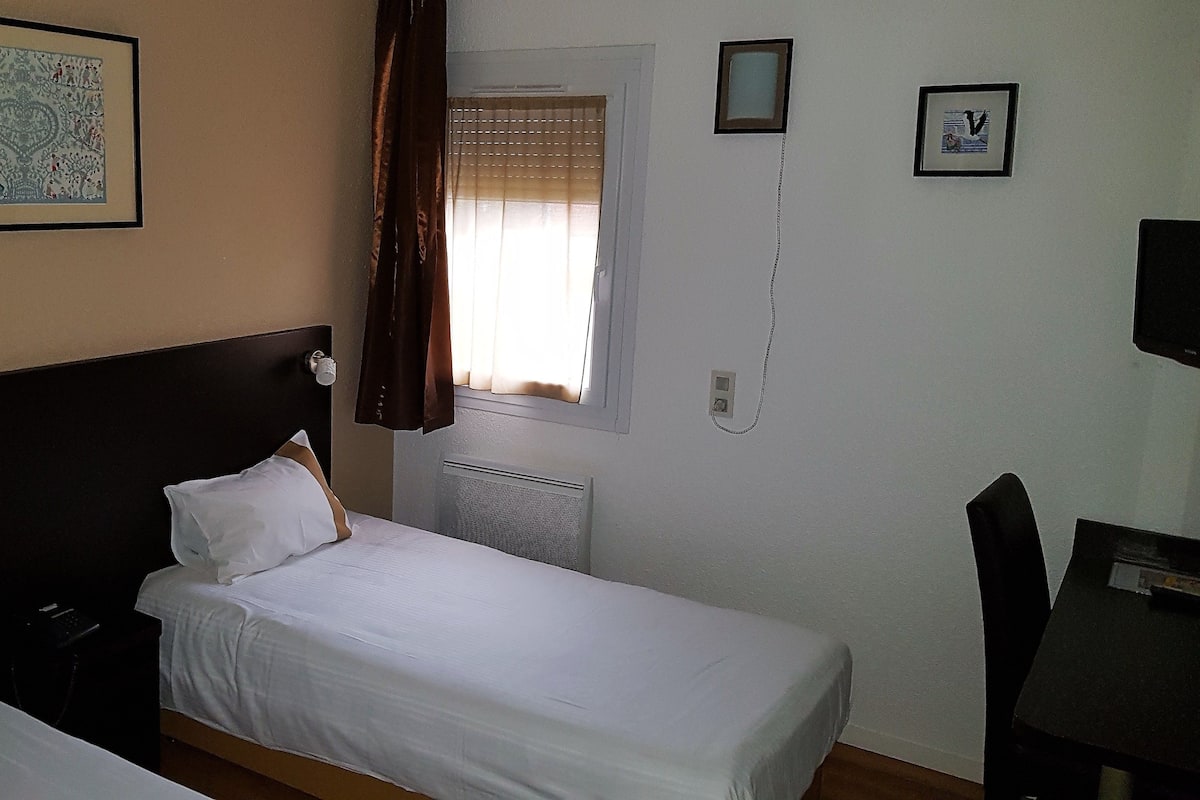 A hotel room with two single beds, a small table, a chair, a window with blinds, and a wall-mounted TV.