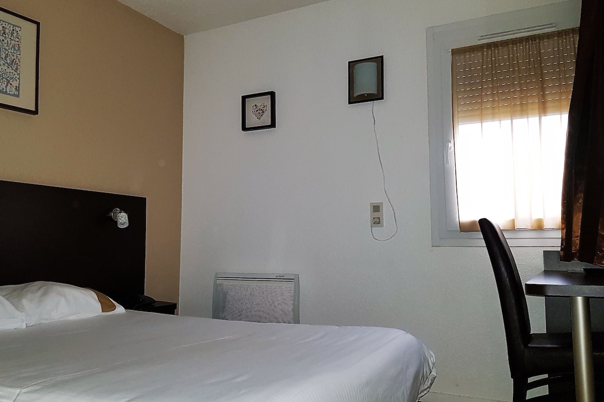 A hotel room with a bed, a desk, a chair, a heater, and a window with curtains.