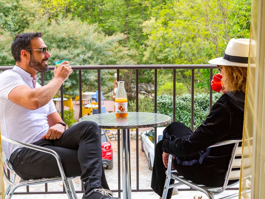 Two people seated at a round table with a bottle of orange juice on it, outdoors with a view of trees and a car in the background.