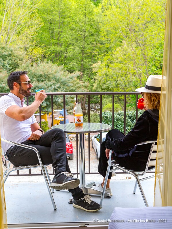 Two people seated at a round table with a bottle of orange juice on it, outdoors with a view of trees and a car in the background.
