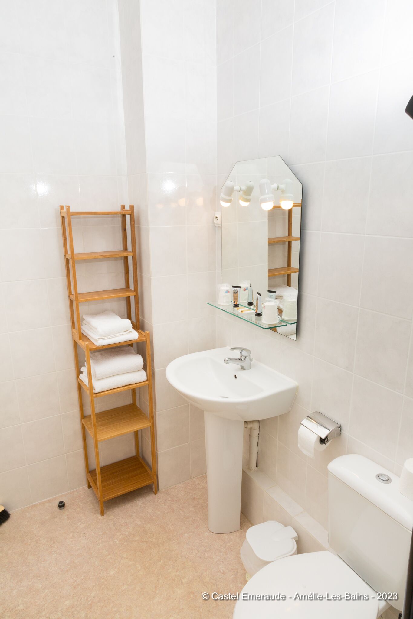 Un baño con un estante de madera que sostiene toallas, un lavamanos blanco, un espejo y un inodoro.
