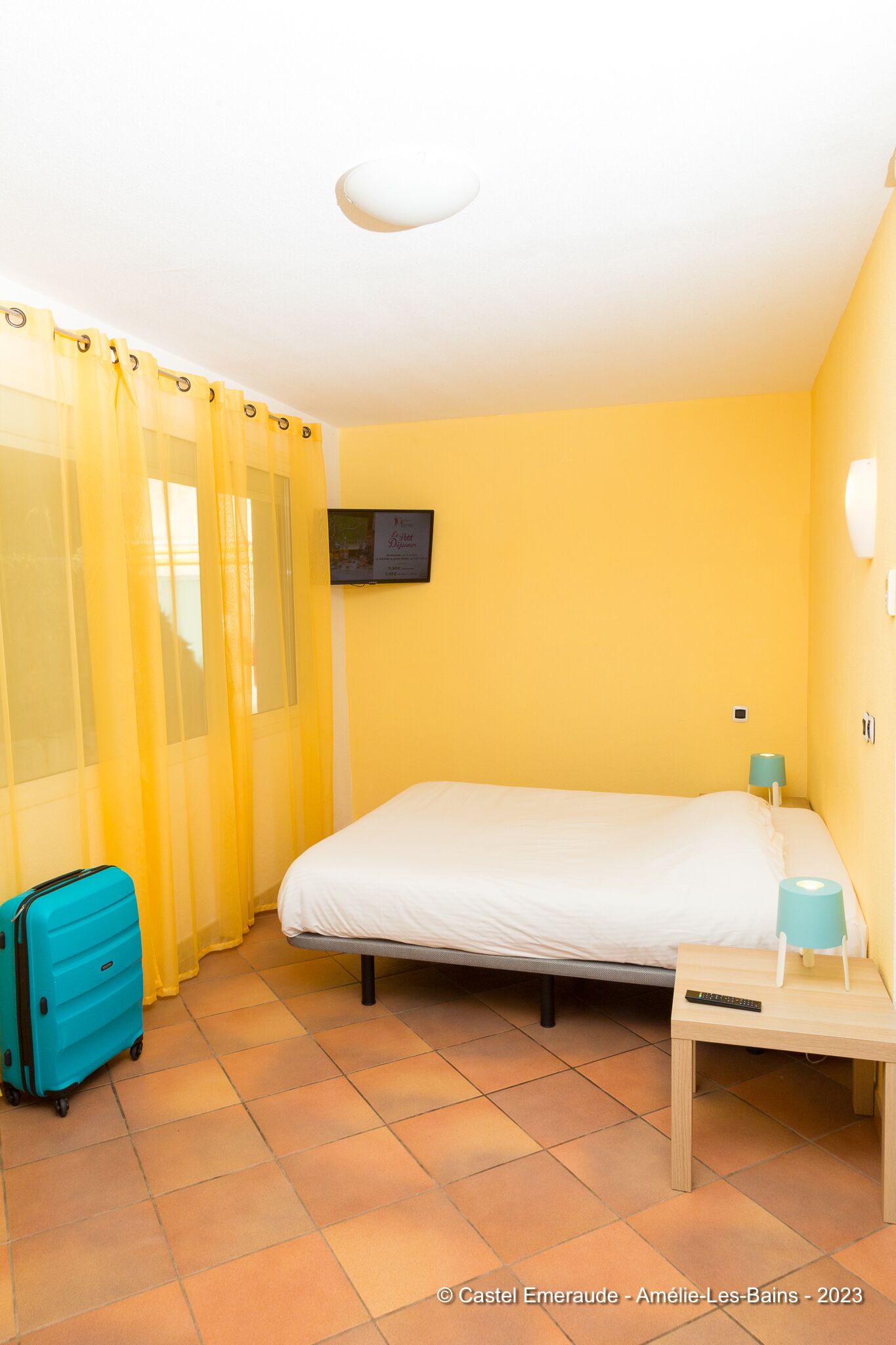 Een hotelkamer met een bed, een klein nachtkastje met lamp, een koffer en een televisie aan de muur.