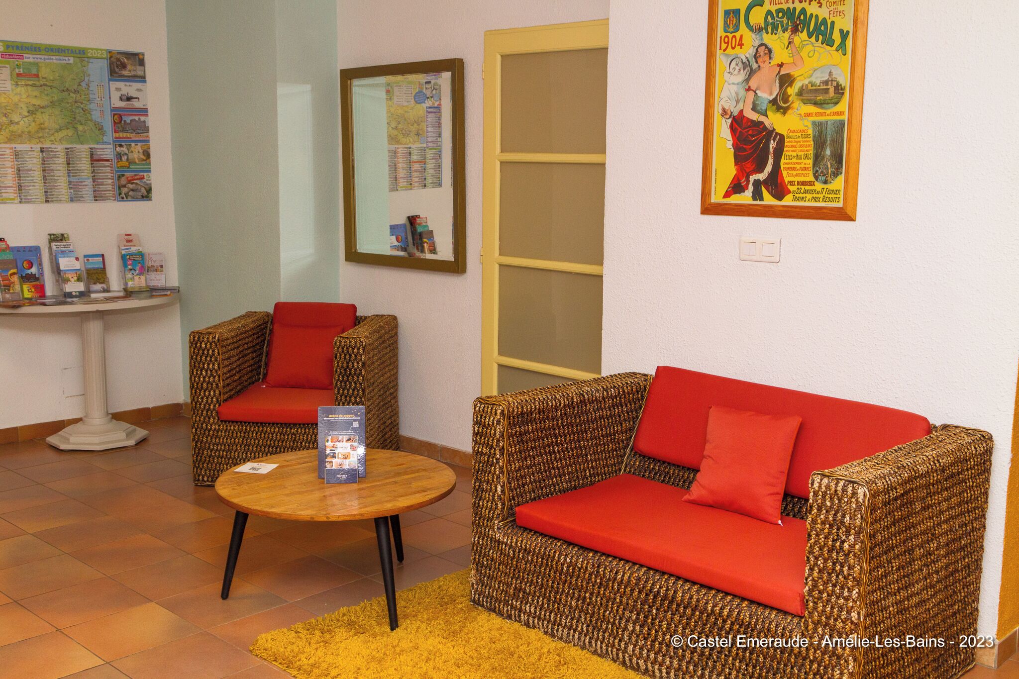 A room with wicker furniture, red cushions, a round wooden table, and a map on the wall.